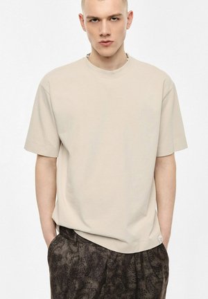 Giovane uomo con capelli corti che indossa una t-shirt beige oversize e pantaloni scuri con fantasia, in piedi con le mani in tasca davanti a uno sfondo bianco.