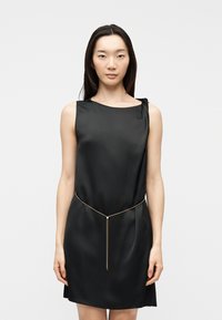 Γυναίκα σε μαύρο σατέν φόρεμα χωρίς μανίκια με λεπτή χρυσή αλυσίδα στη μέση, στέκεται μπροστά σε απλό ανοιχτό γκρι φόντο.