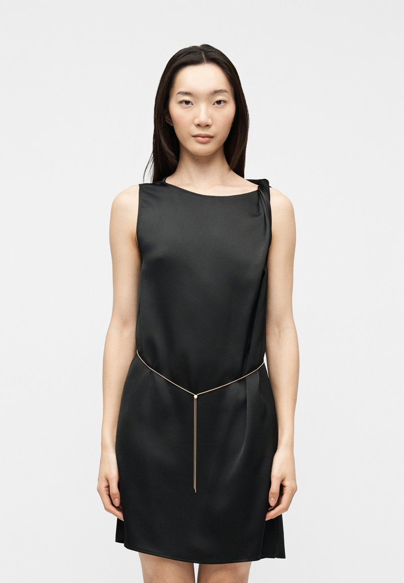 Γυναίκα σε μαύρο σατέν φόρεμα χωρίς μανίκια με λεπτή χρυσή αλυσίδα στη μέση, στέκεται μπροστά σε απλό ανοιχτό γκρι φόντο.