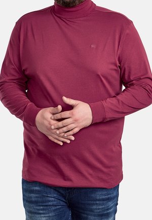 Mann trägt ein langärmeliges burgunderrotes Rollkragenhemd und blaue Jeans, die Hände vor dem Körper verschränkt, vor einem schlichten weißen Hintergrund.