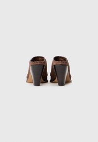 Zapatos de tacón de ante marrón con punta redondeada y costura decorativa a lo largo del borde. Los tacones son de madera oscura y las suelas son de color beige.