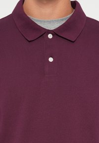 Burgundy polo shirt with a textured knit fabric, a classic collar, three button placket, and a gray undershirt visible underneath.