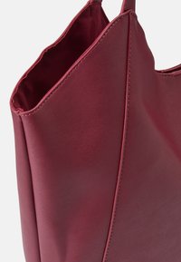 Burgundowa skórzana torba typu tote o gładkiej teksturze, kanciastym designie, otwartym topie i widocznych detalach szycia. Brak akcentów metalowych.