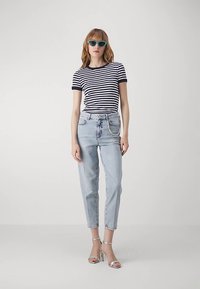 Prugasta mornarsko plava i bijela majica kratkih rukava, svijetloplave traperice s visokim strukom i srebrne sandale s petom; žena nosi tirkizne sunčane naočale.