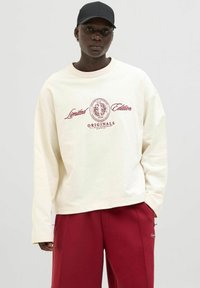 Sudadera de gran tamaño color crema con mangas largas, texto gráfico "Edición Limitada" y "ORIGINALS" en color burdeos. Combinada con pantalones rojos.