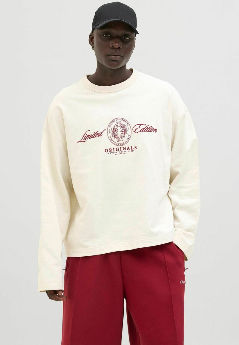 Sudadera de gran tamaño color crema con mangas largas, texto gráfico "Edición Limitada" y "ORIGINALS" en color burdeos. Combinada con pantalones rojos.