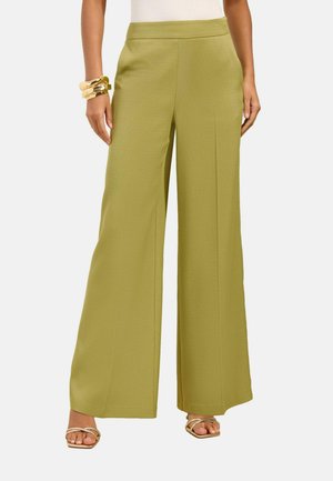 Naine kannab kõrge vöökohaga, laia säärega oliivrohelisi pükse esikokkupressidega, kuldseid käevõrusid ja avatud varvastega kuldseid sandaale valgel taustal.