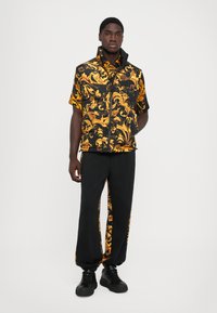 Homme portant une chemise à manches courtes noire et dorée à motifs, avec un gilet assorti et un pantalon noir avec des bandes latérales dorées à motifs.