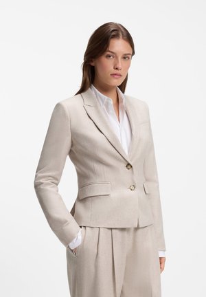 Vrouw draagt een beige op maat gemaakte blazer met twee knopen en bijpassende hoge taille broek, witte overhemd met kraag eronder, poseert met één hand in de zak.