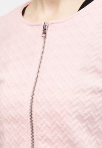 Κοντινό πλάνο υφάσματος σε ανοιχτό ροζ χρώμα με υφή, με ένα ασημί φερμουάρ που τρέχει κάθετα στο κέντρο μπροστά ενός ενδύματος.