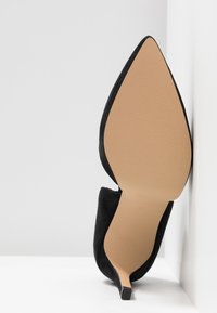 Une chaussure à talon haut en daim noir avec un bout pointu et un talon fin et structuré. La semelle est d'une couleur beige lisse, mettant en valeur une finition texturée.