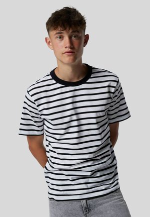 Joven con el cabello corto y rizado, vistiendo una camiseta blanca y negra a rayas y unos jeans gris claro, frente a un fondo gris liso.