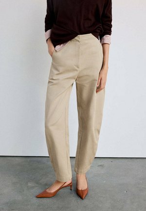 Persona que lleva pantalones beige de talle alto, tacones marrones puntiagudos y una blusa oscura con mangas de camisa a rayas, de pie sobre un suelo gris frente a una pared blanca.