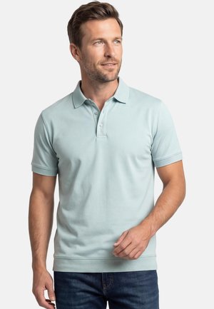 Man draagt een lichtblauw poloshirt met korte mouwen en donkere blauwe spijkerbroek, kijkt iets naar rechts tegen een effen achtergrond.