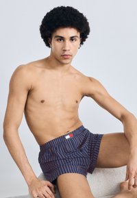 Μπόξερ με ρίγες σε μπλε ναυτικό και κόκκινο χρώμα, με ελαστική μέση και ταμπέλα με λογότυπο. Το ύφασμα φαίνεται μαλακό και ελαφρύ, πέφτοντας άνετα.