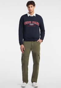 Sudadera de marina con el logo "GUESS JEANS 1981" en rojo, combinada con pantalones cargo verdes oliva que tienen múltiples bolsillos, y zapatillas blancas.