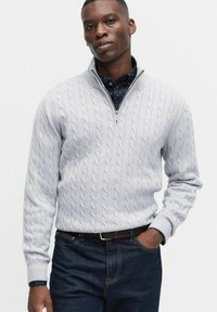 Jasnoszary sweter w splot kablowy z kołnierzem na zamek, noszony na tle ciemnej koszuli w kropki, zestawiony z ciemnoniebieskimi dżinsami.