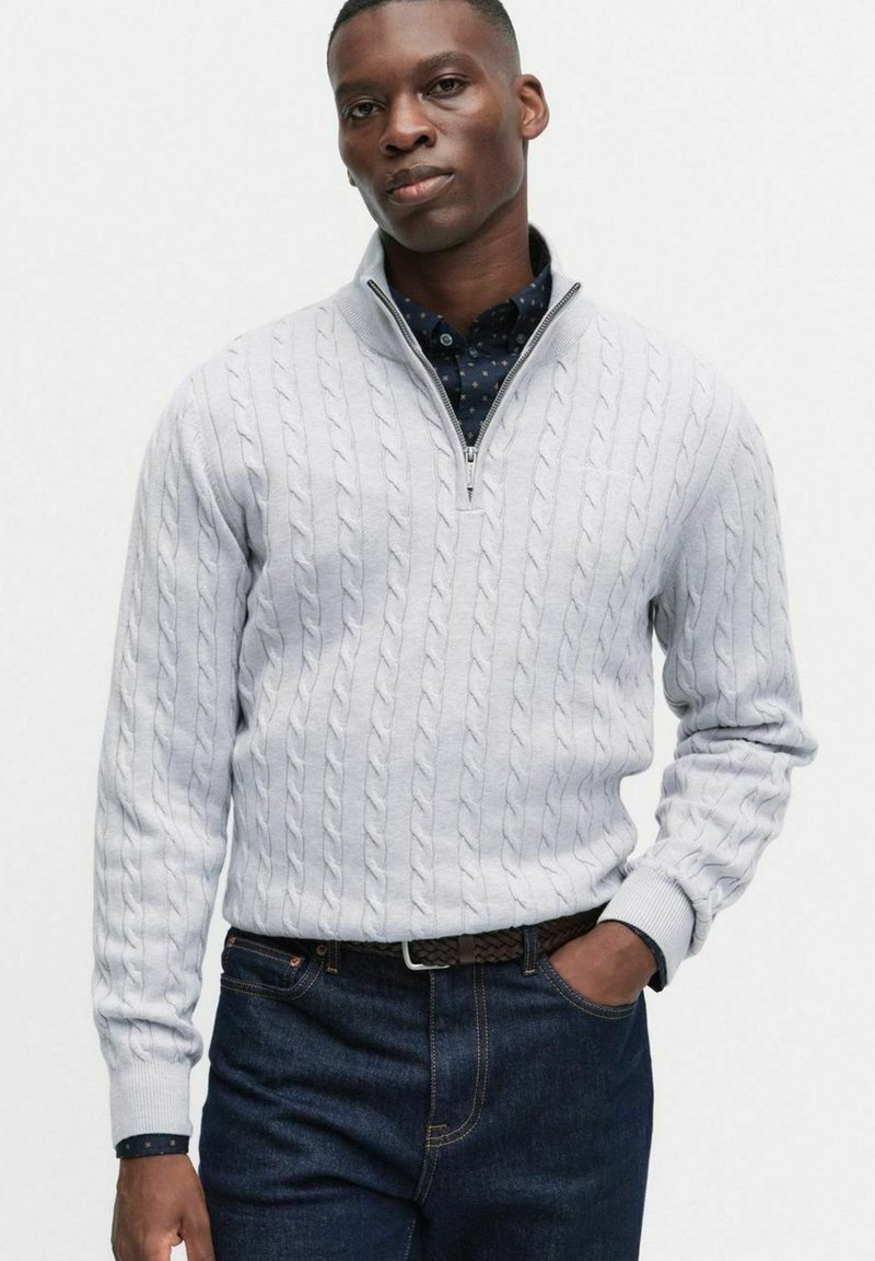 Jasnoszary sweter w splot kablowy z kołnierzem na zamek, noszony na tle ciemnej koszuli w kropki, zestawiony z ciemnoniebieskimi dżinsami.