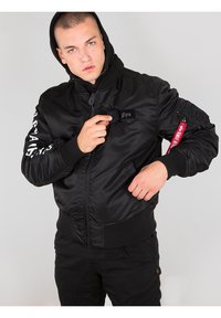 Čierna bomber bunda so zipsovým uzáverom, rebrovanými manžetami a kapucňou. Obsahuje logo nášivku a červenú visačku na rukáve. Hladký povrch.