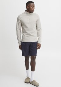 Light beige long-sleeve top with a zip collar, paired with dark navy shorts, white socks, and beige slip-on shoes. Simple, casual design.