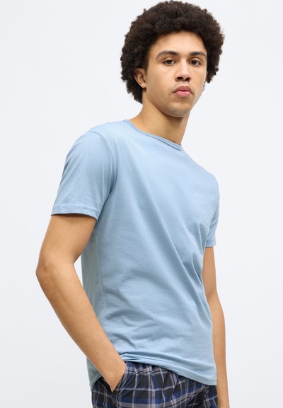 Tricou din bumbac albastru deschis, cu guler rotund și mâneci scurte, cu un logo discret pe piept. Asortat cu pantaloni scurți în carouri.