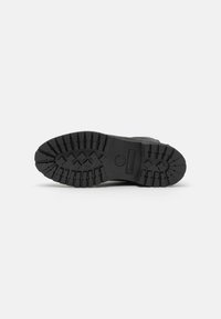 Suela de bota de goma negra con un patrón de surcos profundos, que presenta una impresión de logo. Superficie texturizada para agarre, bordes curvados para contorno.