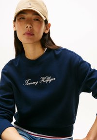 Felpa blu navy con ricamo bianco "Tommy Hilfiger", polsini e orlo a coste. Abbinata a un cappellino blu chiaro.