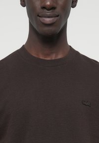 Καφέ μπλούζα με στρογγυλή λαιμόκοψη από υφασμένο βαμβάκι, με το μαύρο λογότυπο Lacoste κεντημένο στην αριστερή πλευρά του στήθους.