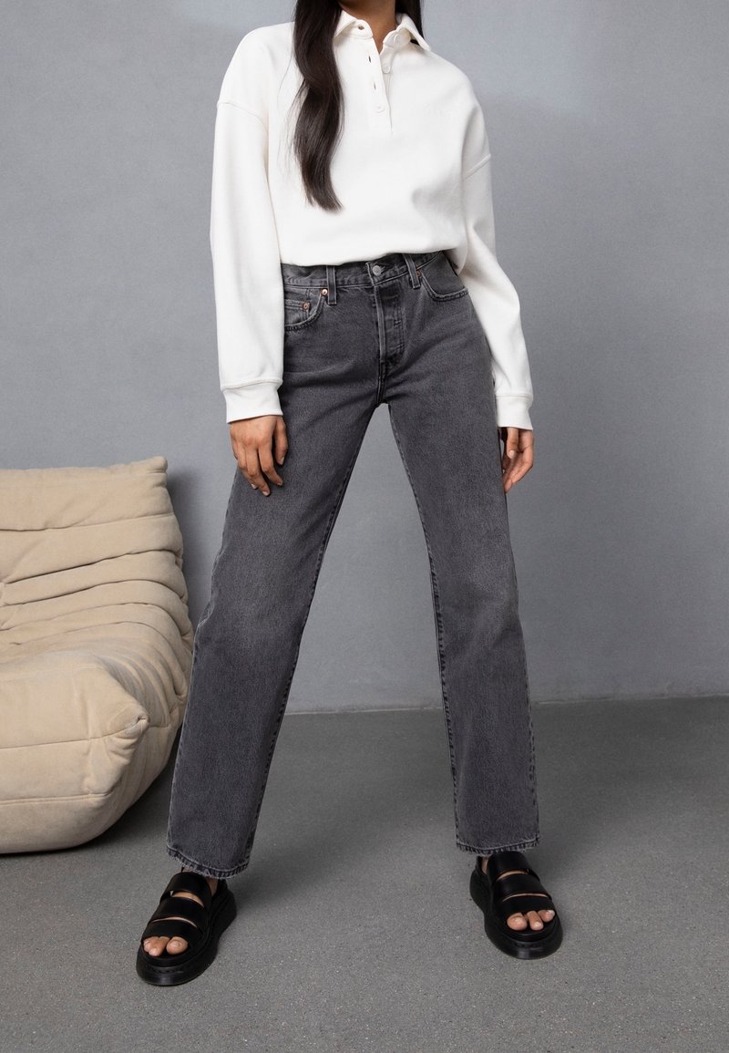 Personne debout en jean noir délavé taille haute, polo blanc à manches longues et sandales noires à bout ouvert sur fond gris.