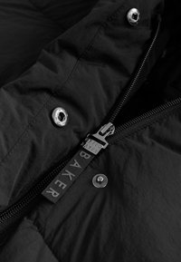 Schwarze Pufferjacken-Innenseite mit einem silbernen Reißverschluss und einem schwarzen Tab mit der Aufschrift "Ted Baker" sowie metallischen Druckknöpfen, gestaltet mit einer glatten Textur.