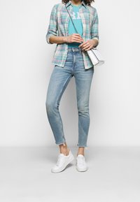 Mladá žena oblečená v světle modrých skinny jeans, bílých teniskách, tyrkysovém tričku s potiskem, otevřené kostkované košili a nese malou bílou kabelku na rameni.