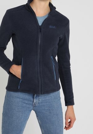 Persoon draagt een navyblauwe fleecejas met rits en geritselde zakken, gecombineerd met lichtblauwe spijkerbroek tegen een effen achtergrond.