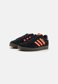 Mustat mokkanahkaiset tennarit, joissa on kolme oranssia raitaa ja pintamateriaalina poimuttunut kuviointi. Kenkien pohja ja kantapään yksityiskohdat ovat ruskeaa kumia.