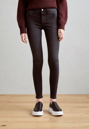 Osoba ubrana w ciemne obcisłe dżinsy, czarne platformowe buty wsuwane z białymi podeszwami oraz bordowy sweter, stojąca na drewnianej podłodze.