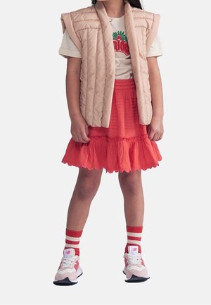 Enfant portant un gilet matelassé beige, un t-shirt graphique blanc, une jupe rouge à volants, des chaussettes rayées rouges et blanches, et des baskets roses, debout.