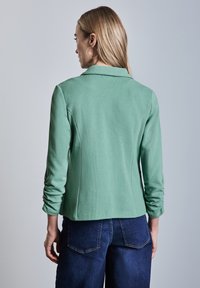 Mintgroene, fitted jas met een zachte textuur, lange mouwen en een geknipte kraag. Gepaard met donkerblauwe spijkerbroek.