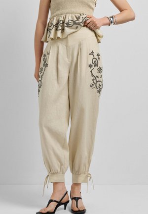 Beige broek met hoge taille en zwarte bloemrijke borduursels op de dijen, taps toelopende enkels met strikjes, gedragen met zwarte sandalen met hoge hak en bandjes.
