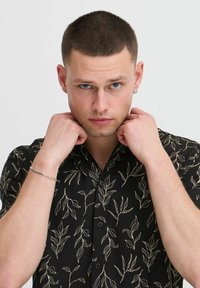 Chemise noire à manches courtes avec un motif de feuilles vertes et beige. Elle présente un col et une fermeture à boutons sur le devant, maintenue par des mains au niveau du col.