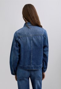 Denimjakke i mørkeblå med krave, lange ærmer og en let cropped pasform. Har synlige syninger og et klassisk rygstykke.