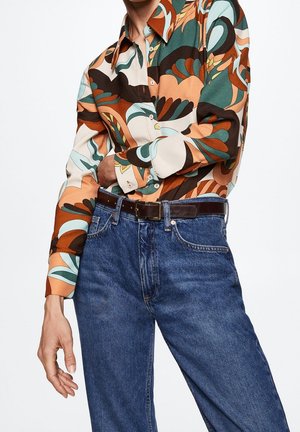 Person wearing a colorful, abstract patterned long-sleeve shirt tucked into high-waisted blue jeans with a dark brown belt.