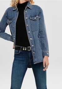 Leren spijkerjack in lichtblauw met zilveren knopen, twee borstzakken, een kraag en lange mouwen, gedragen over een zwarte top en jeans.