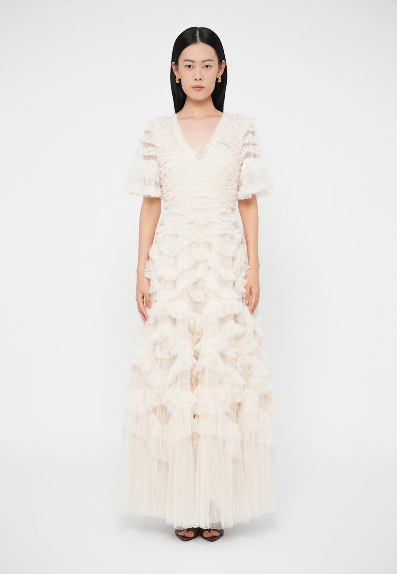 Kobieta w kremowej sukni sięgającej do podłogi z warstwami falbanek i przejrzystymi krótkimi rękawami, stojąca na tle jednolitego białego tła.