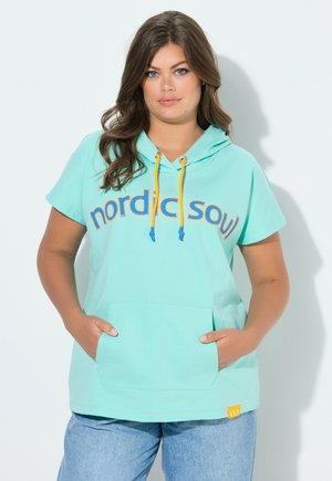 Frau mit braunen Haaren trägt einen mintgrünen Kurzarm-Hoodie mit gelben Kordeln und dem Schriftzug „nordic soul“, Hände in den Taschen, vor weißem Hintergrund.