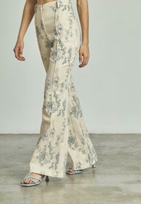 Vrouw in crèmekleurige, bloempatroon wijdgerande broek en lichtblauwe, gespen met hakken, staande op een gladde betonnen vloer.