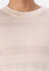 Svetlo bež pletena pulover s teksturiranimi horizontalnimi črtami in rebrastim okroglim izrezom. Material se zdi mehak in zračen.