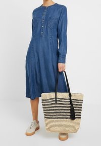 Denimová šaty s dlouhými rukávy, zapínáním na knoflíky a dvěma náprsními kapsami. Drží pletenou slaměnou tašku s černými pruhy a třásní.