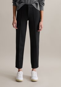 Schwarze maßgeschneiderte Hose mit glatter Textur, vorne mit Falten, einem strukturierten Bund und schmalen Beinöffnungen, kombiniert mit weißen Sneakers.