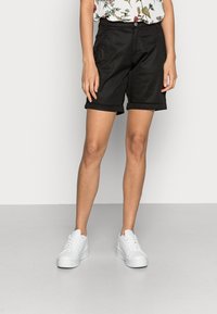 Svarta shorts med skräddarsydd passform, vikta fållar och sidofickor. Tillsammans med vita sneakers, mot en enkel bakgrund.