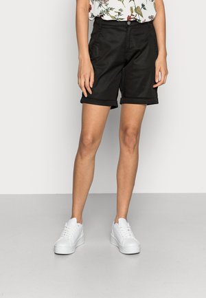 Pantalones cortos negros con un corte entallado, puños doblados y bolsillos laterales. Combinados con zapatillas blancas, sobre un fondo liso.