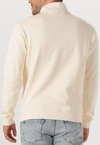 Crème-kleurige sweatshirt met hoge kraag, ribgebreide manchet en zoom, zachte textuur en een klein logo-detail op de achterkant van de nek.